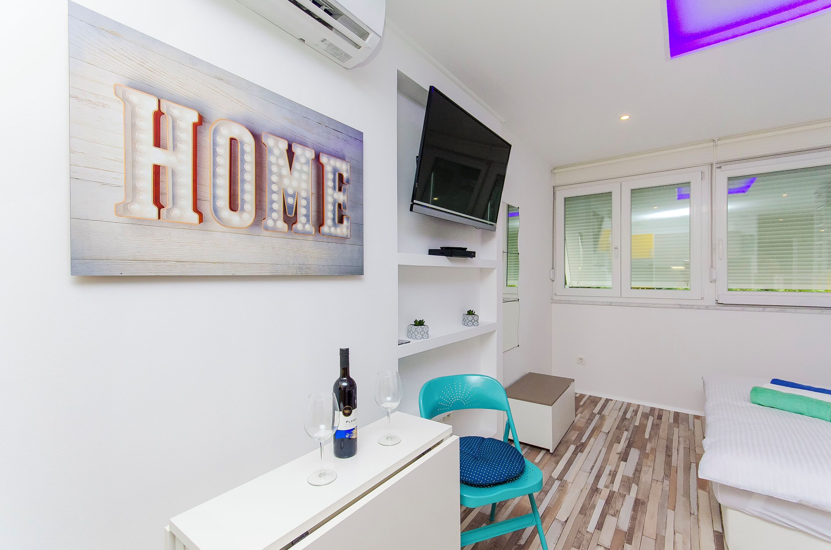 Une chambre d’hôtel moderne avec un panneau indiquant « HOME », un téléviseur à écran plat, une petite table avec une bouteille et des verres, un fauteuil turquoise et un lit avec un oreiller vert.