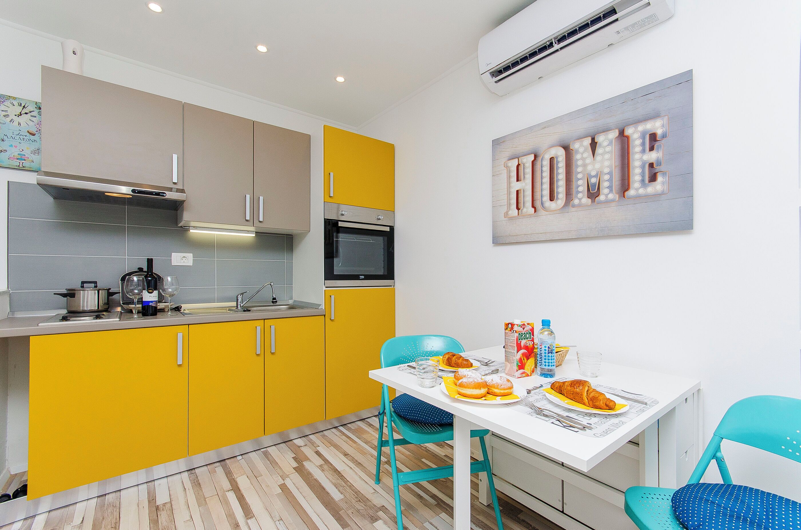 A compact kitchen with yellow cabinets, a white dining table, and a "HOME" sign on the wall.
