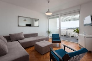 A modern living room with a sectional sofa, ottoman, and chairs, a large mirror, and a view through a sliding glass door to an outdoor area with a table and chairs. - Apartment Town and Beach (Makarska)