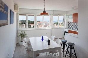 A modern kitchen with white cabinets, a dining table with chairs, and a view of the city through large windows. - Apartment Town and Beach (Makarska)