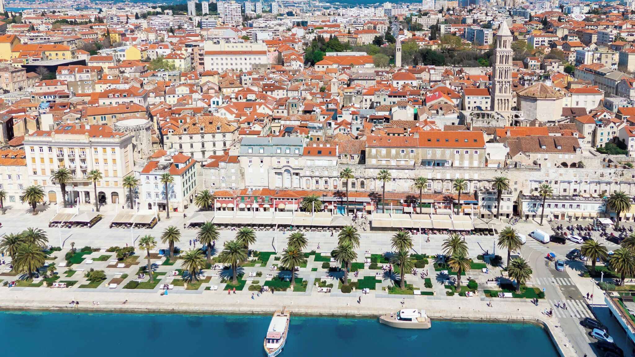A coastal cityscape with historic buildings, a marina, and a promenade with palm trees.