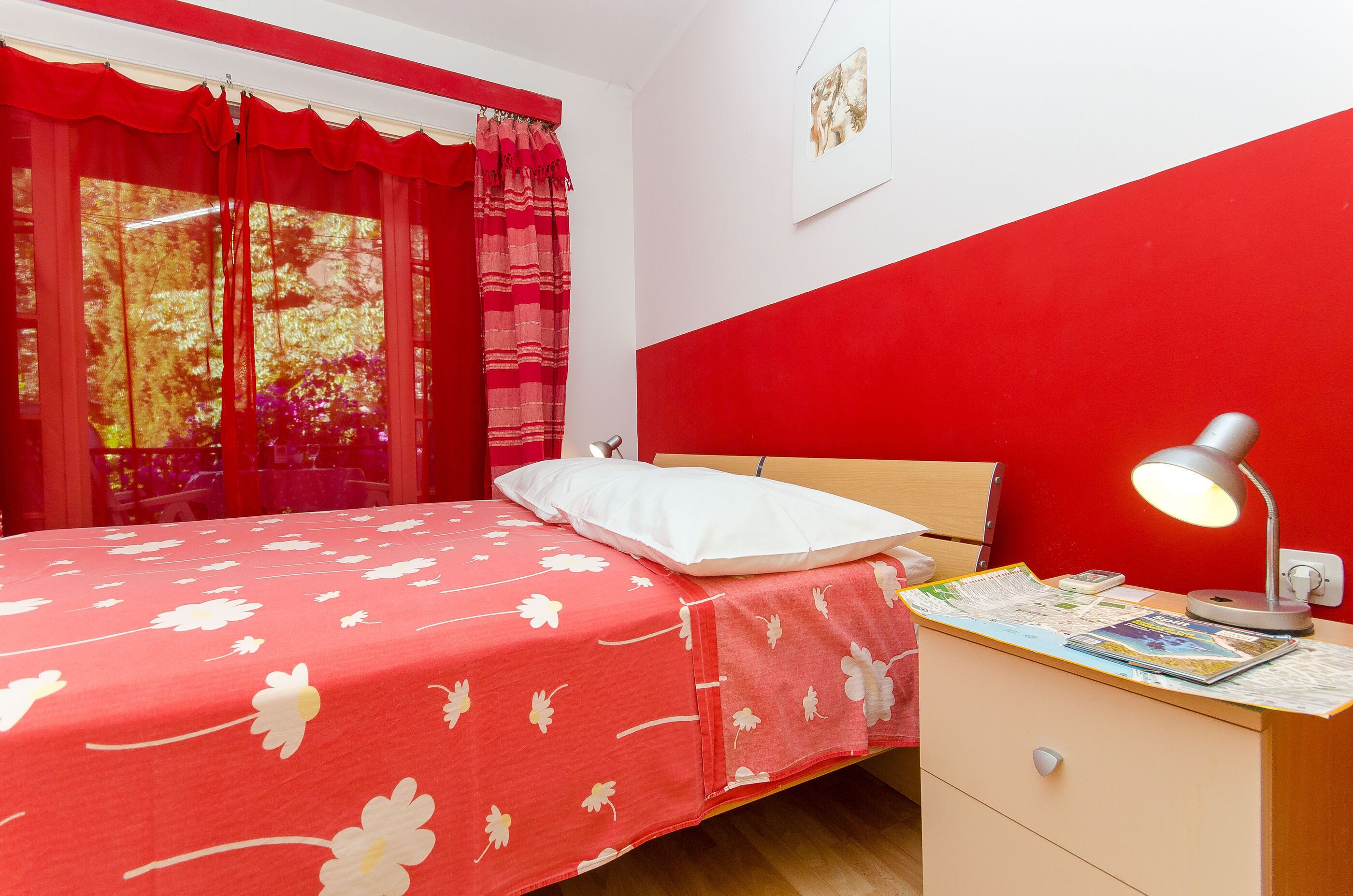 A room with a bed, a nightstand with a lamp, a window with curtains, and a red wall.