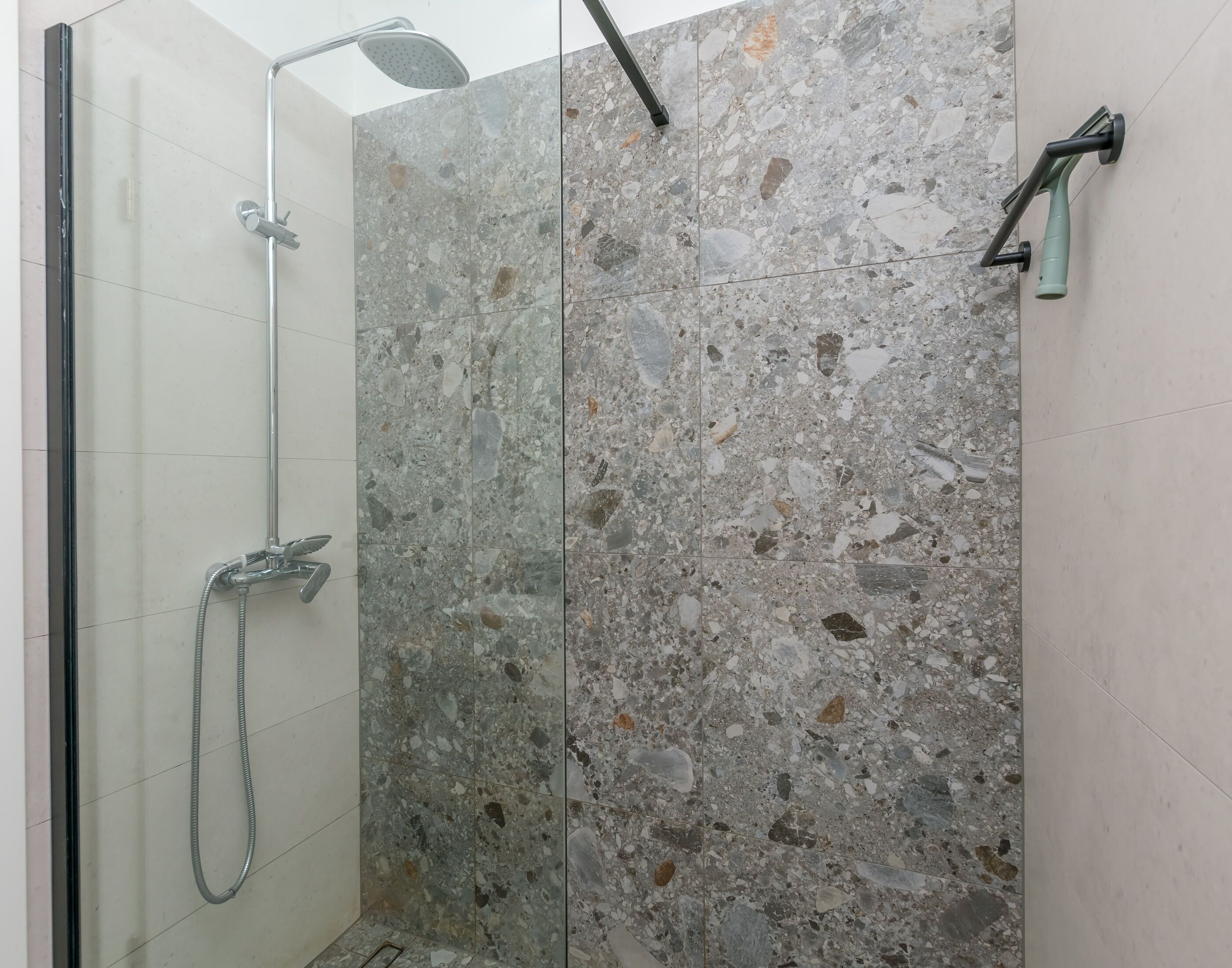 A shower with a handheld showerhead and a wall-mounted faucet, a glass door, and a tiled wall with a speckled pattern.