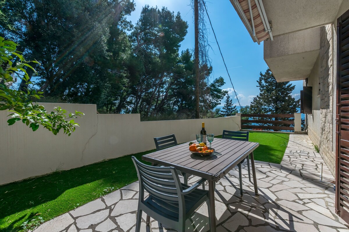 A patio with a table set for two, surrounded by chairs, a small garden, and a view of trees.