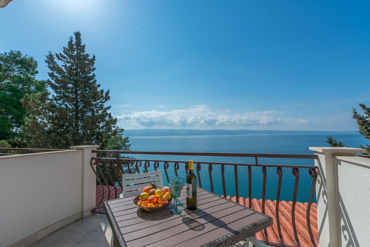 A balcony with a table, a bowl of fruit, and a bottle of wine.