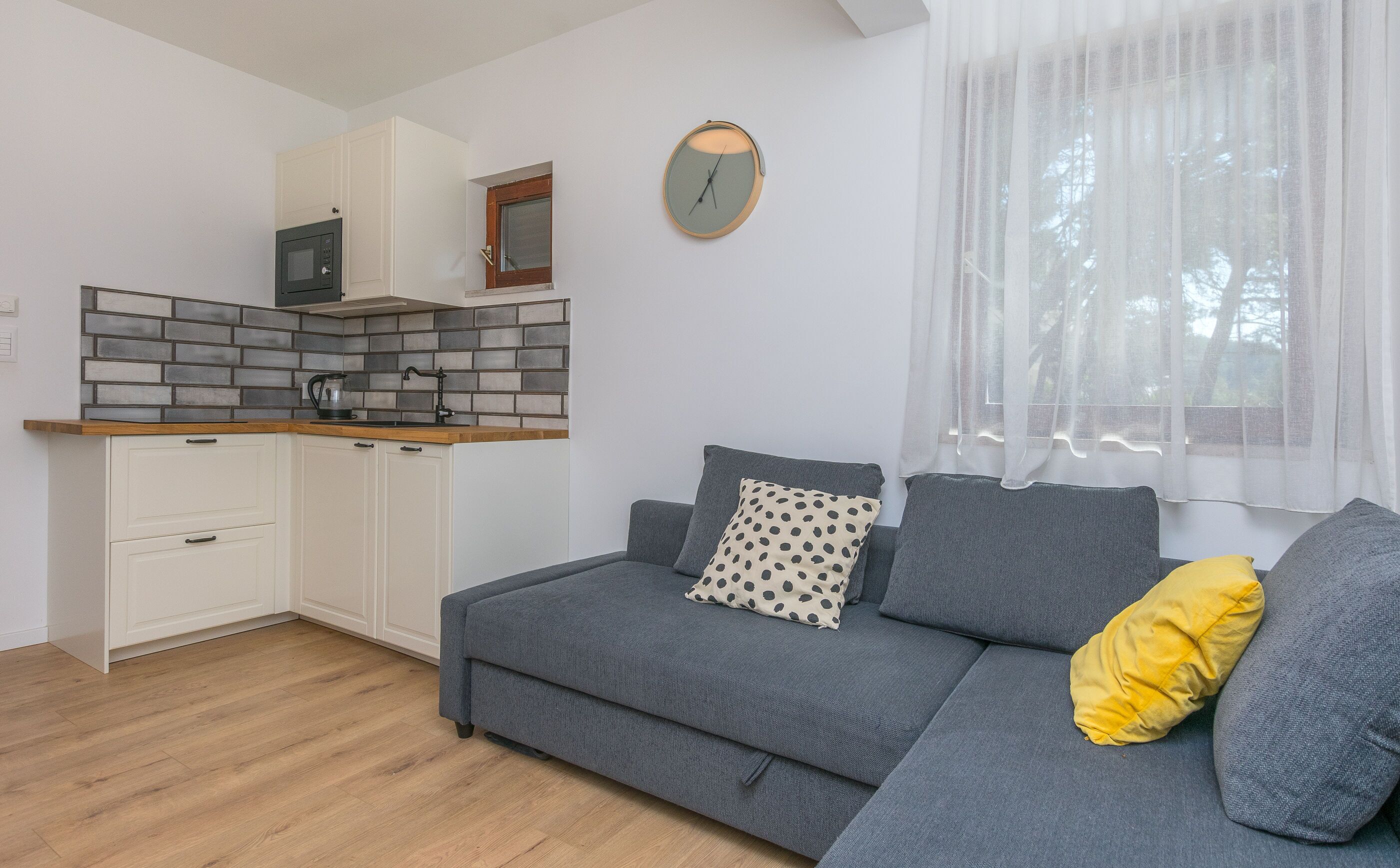A modern living space with a kitchenette, a grey sofa with cushions, and a window with sheer curtains.
