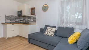 Un espacio habitable moderno con kitchenette, un sofá gris con cojines y una ventana con cortinas traslúcidas.