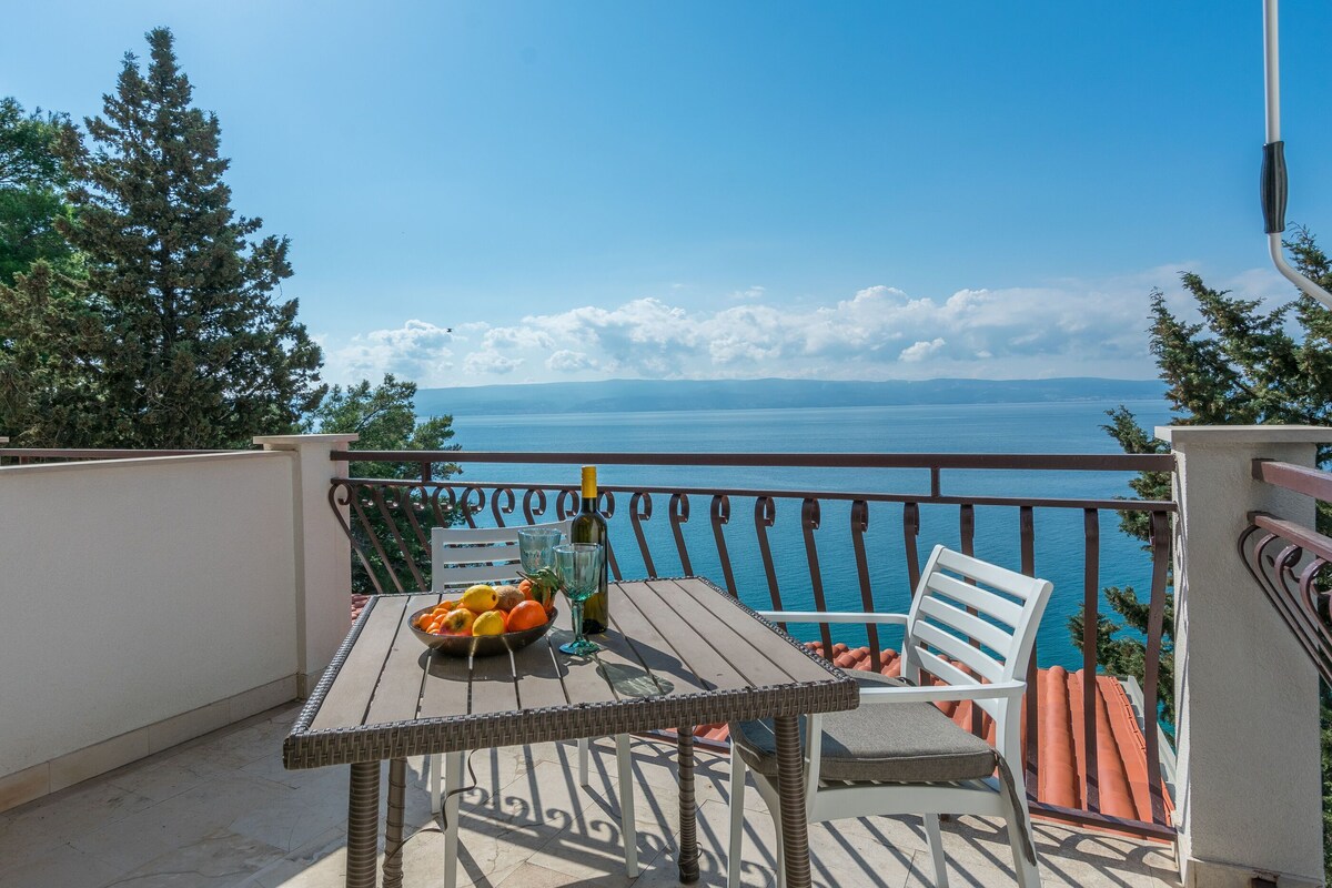 A balcony with a table, chairs, a bottle, and a bowl of fruit.
