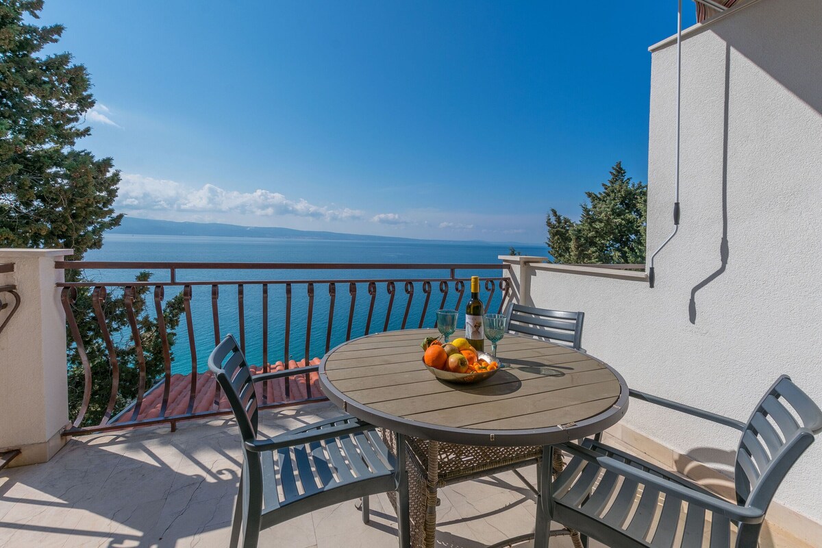 A round table with a fruit bowl, two chairs, and a bottle on a balcony overlooking a body of water.