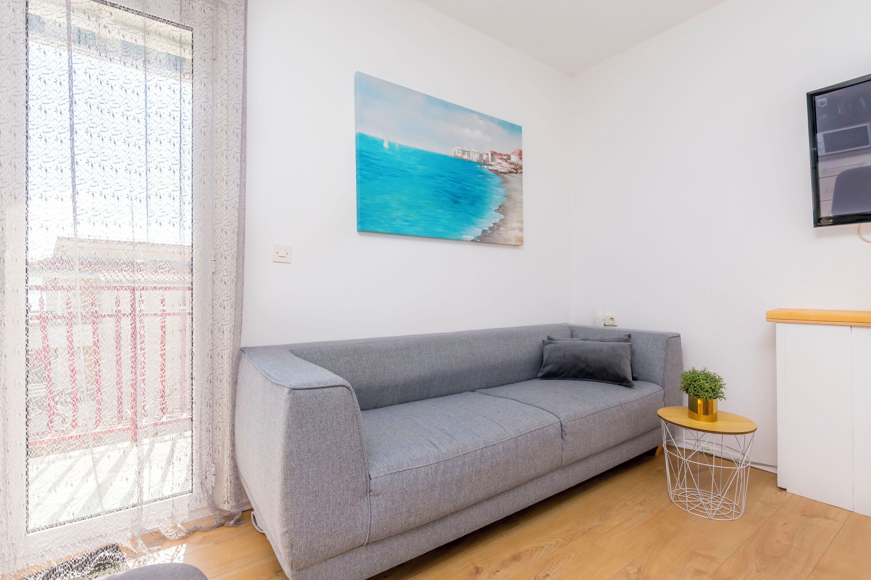 Un salón con un sofá gris, una mesita redonda con una planta, un cuadro de una escena costera y una ventana con cortinas de encaje.