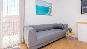 A living room with a grey sofa, a small round table with a plant, a painting of a coastal scene, and a window with lace curtains. - Apartment Jadran (Omis)