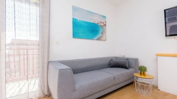 Un salón con un sofá gris, una mesita redonda con una planta, un cuadro de una escena costera y una ventana con cortinas de encaje.