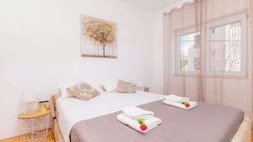 Una cama doble con una colcha rosa, dos almohadas blancas, una mesita de noche con una lámpara, y un cuadro de árboles en la pared.