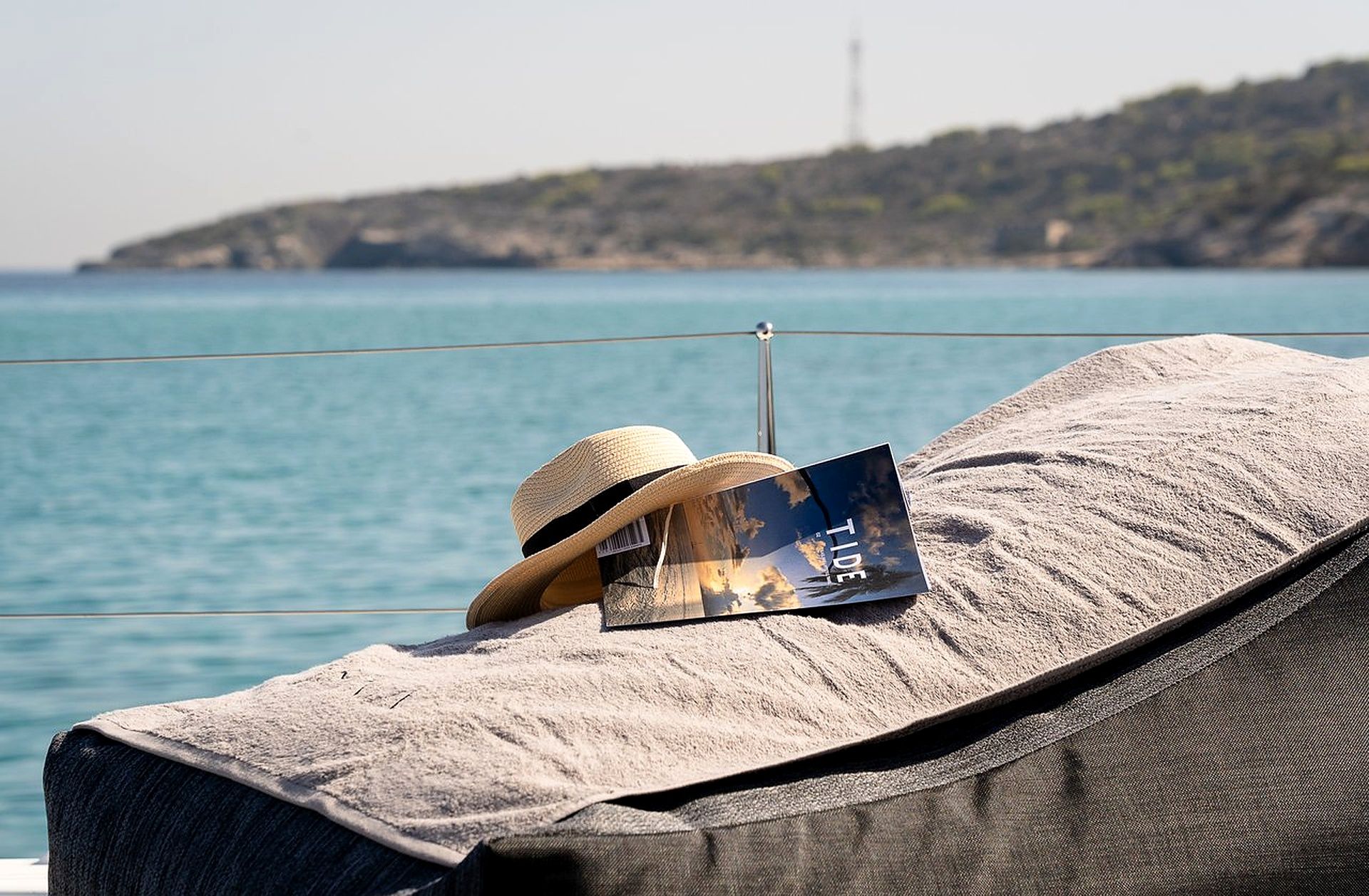 A beach chair with a straw hat and a magazine on a towel, overlooking a calm sea and a hilly landscape.