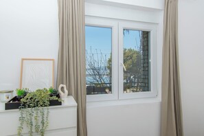 Ein Zimmer mit Fenster, Vorhängen, einer Pflanze, einem gerahmten Bild und einem kleinen Regal mit Gegenständen.