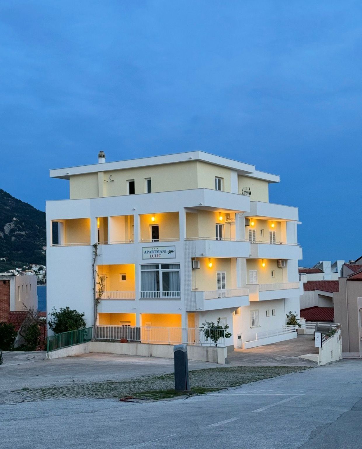 Un edificio de varios pisos con balcones, un letrero que dice 'APARTMANTI LULIĆ' y vistas del entorno.