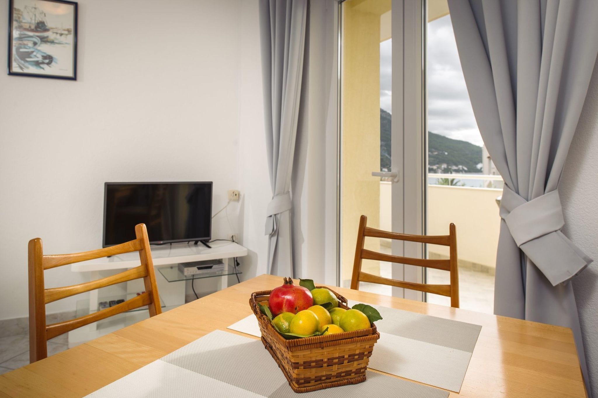 Uma mesa de jantar com uma cesta de frutas, duas cadeiras de madeira, uma televisão e vista do exterior através de uma janela com cortinas.