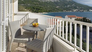 Een balkon met rieten meubels, een tafel met een fruitschaal, en uitzicht op een kustgebied met bergen.