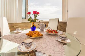 Uma mesa de jantar com tampo de vidro, cadeiras brancas, um vaso com flores vermelhas, uma chaleira, um prato de croissants, uma tigela de morangos e duas xícaras com pires.
