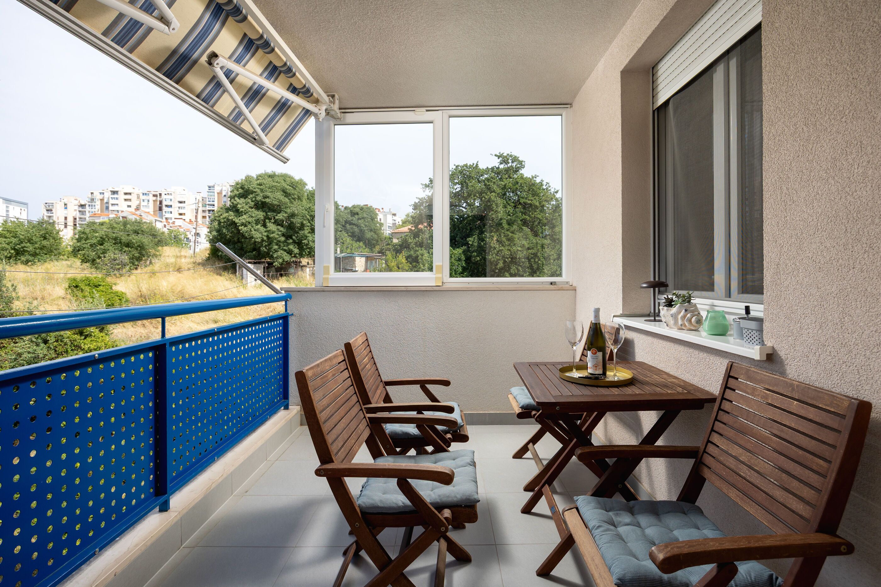 A balcony with a table and chairs, a window with a view of buildings and trees, and a railing.