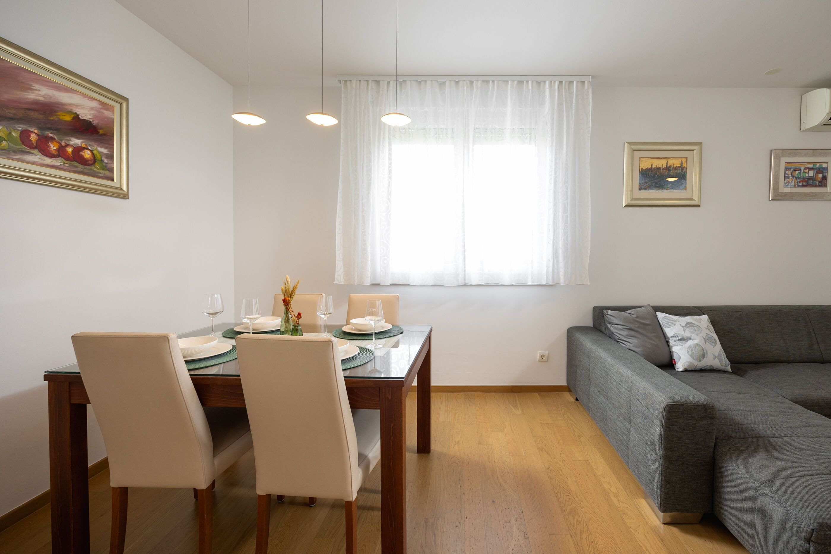 A dining area with a table set for four, a grey sofa, and two framed artworks on the wall.