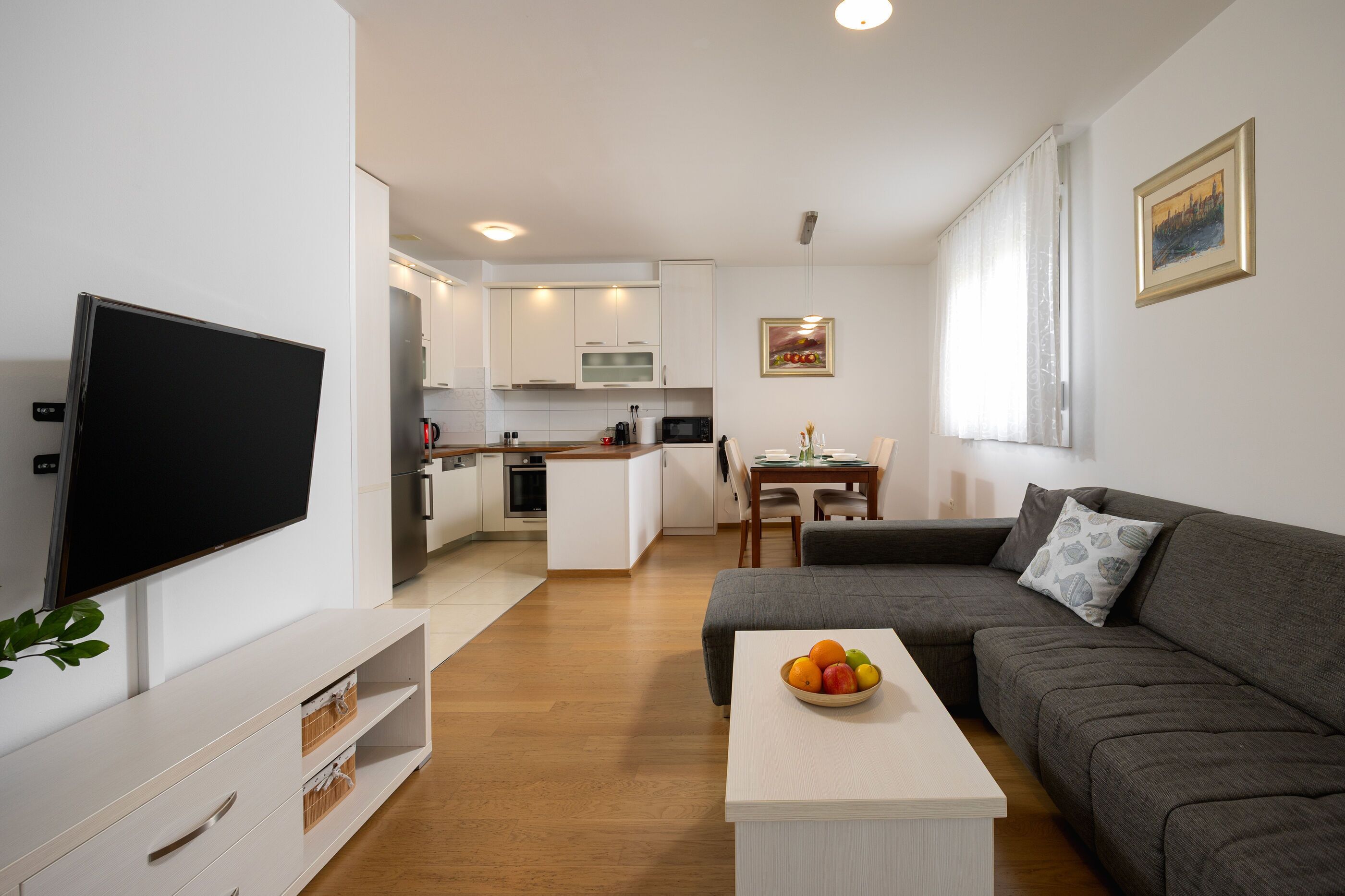 A modern living room with a flat-screen TV, a grey sofa, a coffee table with fruit, and a dining area with a table and chairs.