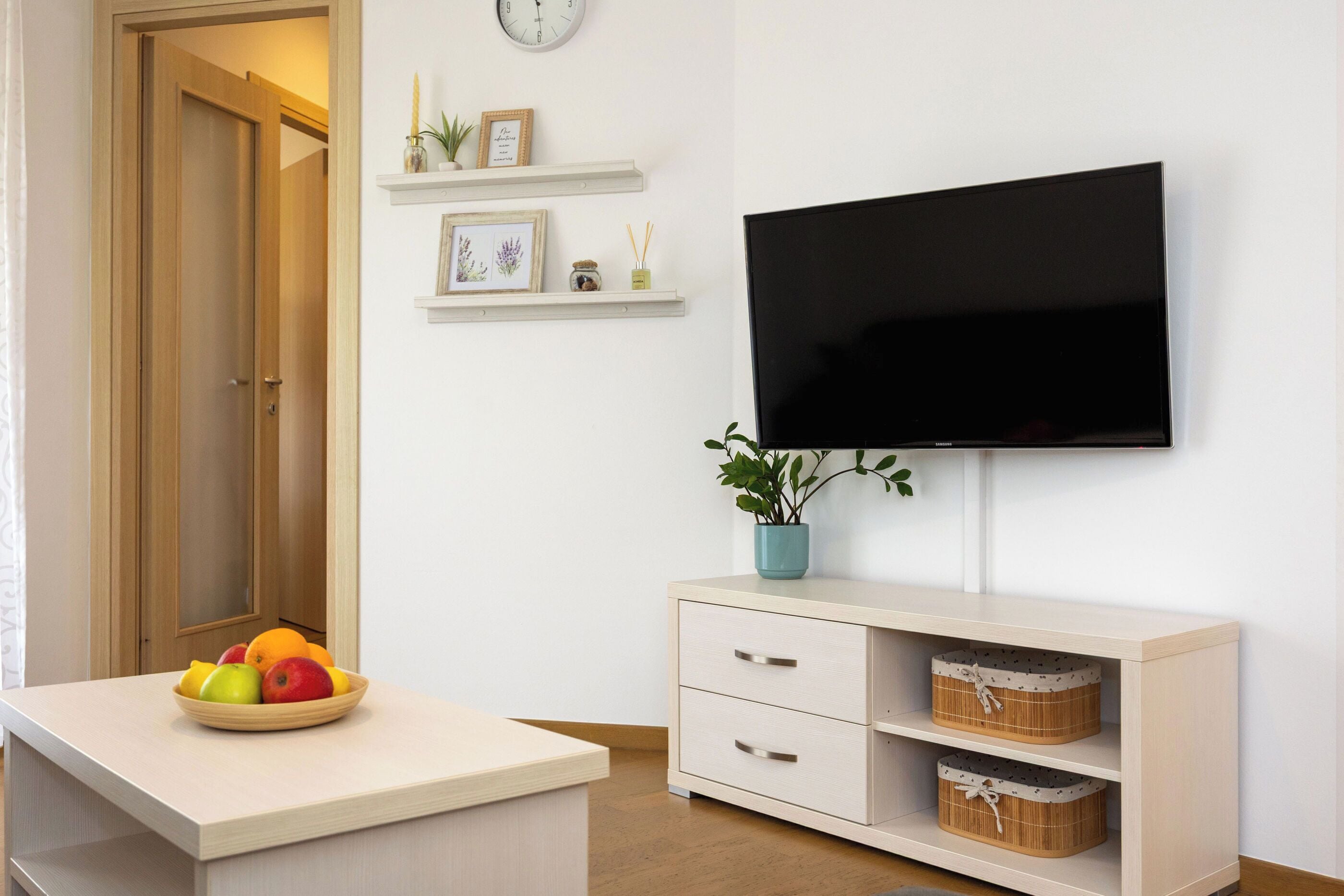 Una sala de estar moderna con un televisor de pantalla plana montado en la pared, un mueble de madera con cajones, una mesita con un cuenco de fruta y una planta en maceta.