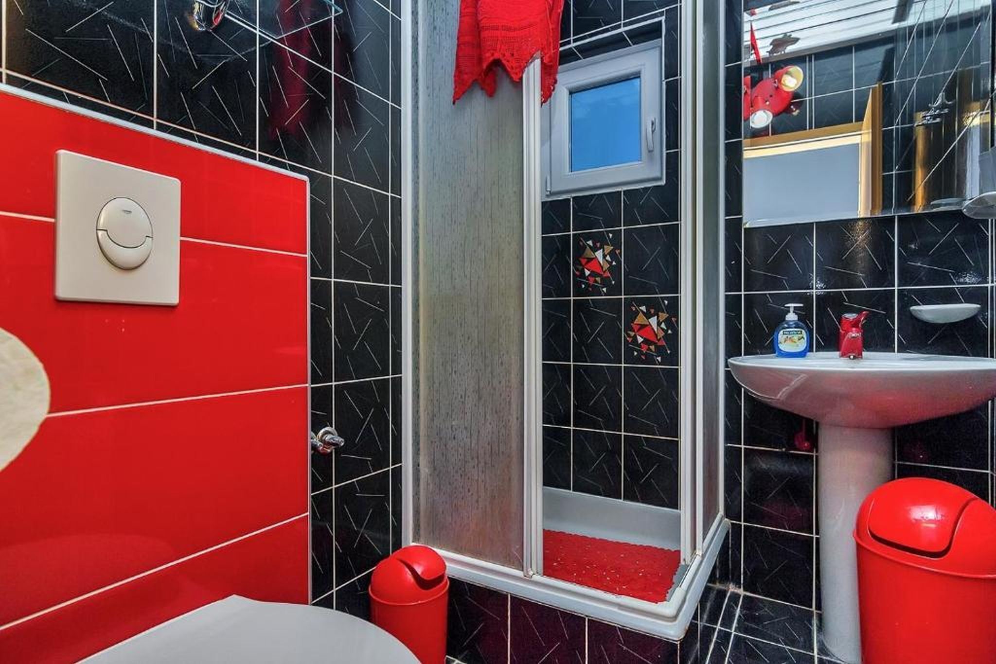 A bathroom with black and red tiled walls, a white sink, and a red trash bin.