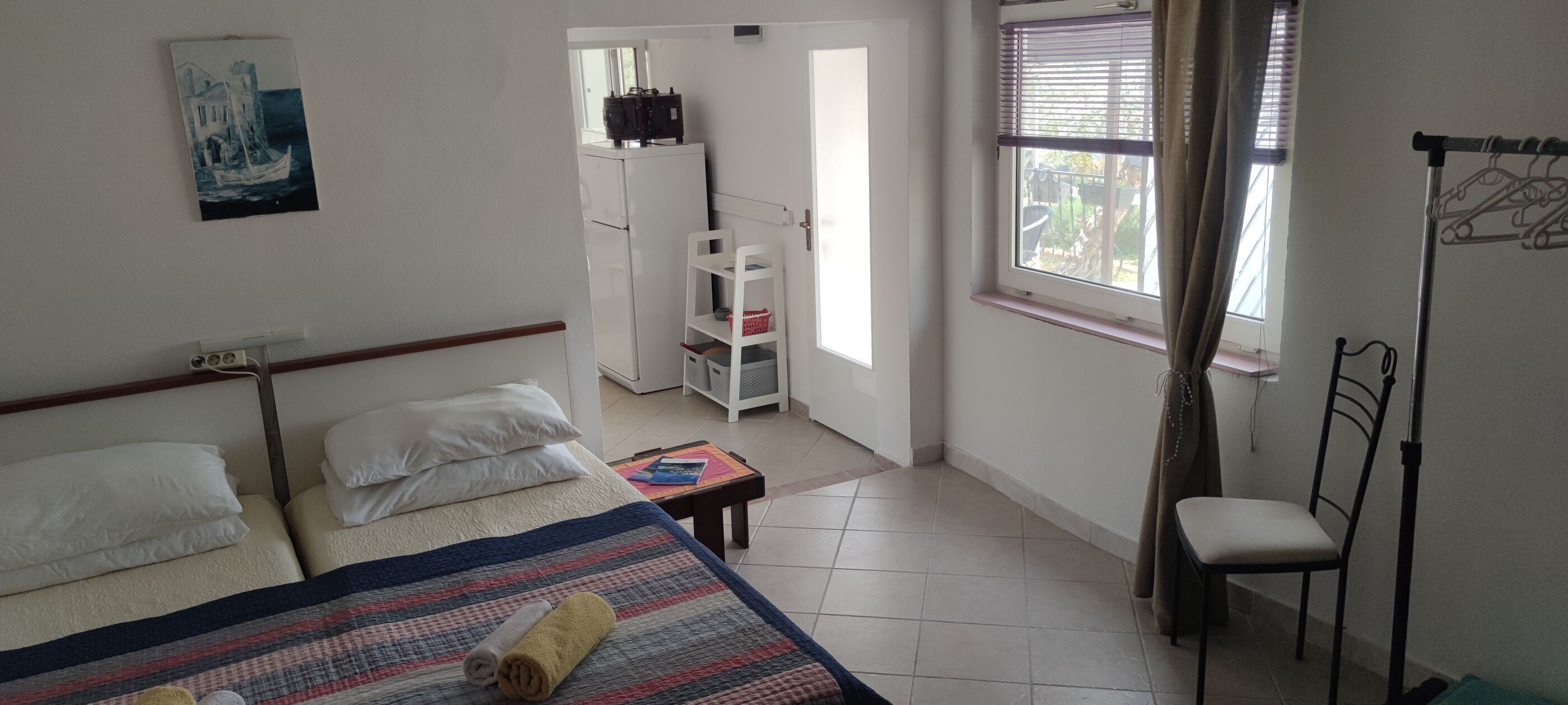 Een slaapkamer met een bed, kussens, een opgerolde handdoek, een tafeltje, een stoel, een kast en een raam met jaloezieën.