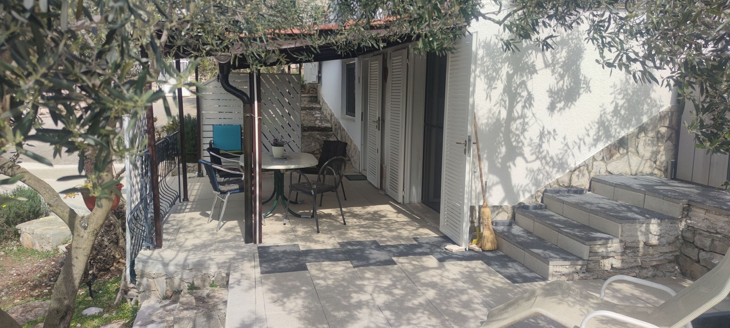 Een overdekt terras met een tafel, stoelen, een bezem en een trap naar een gebouw.