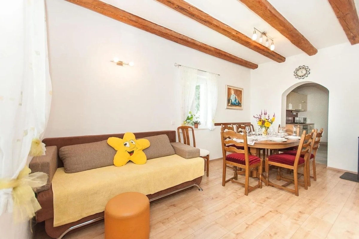A cozy living room with a sofa, a dining table set for four, wooden beams on the ceiling, and a small stool.