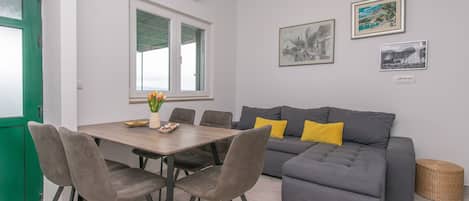 Een moderne woonkamer met een grijze bank, twee gele kussens, een eettafel met vier stoelen en twee ingelijste foto's aan de muur.