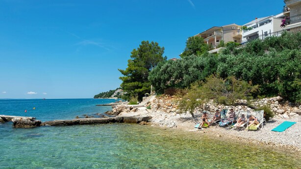 A coastal beach with clear water, rocky shore, and buildings in the background. - Apartment Garden by the Sea (Celina)