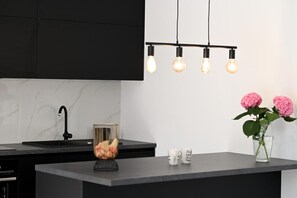 A modern kitchen with a black countertop, a marble backsplash, a black faucet, a fruit bowl, a vase with flowers, and hanging light bulbs. - Villa Serenity With Heated Pool (Donji Prolozac)