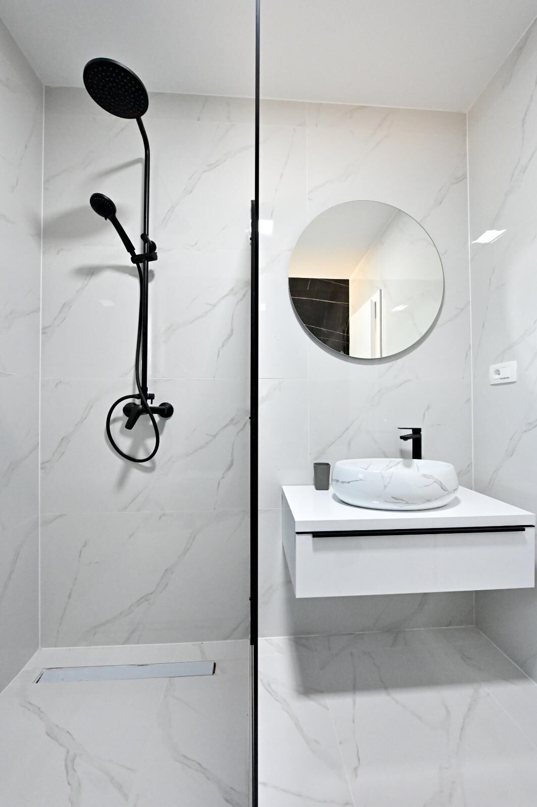 A modern bathroom with a shower, a round mirror, and a white sink with a marble countertop.