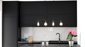 Et moderne køkken med en sort bordplade, et marmorbakpanel, en hængende lampe med fire pærer, en vase med lyserøde blomster og to krus med tekst.