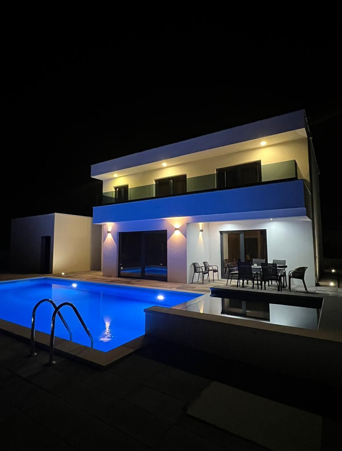 A modern two-story house with a swimming pool, outdoor seating, and a well-lit exterior.