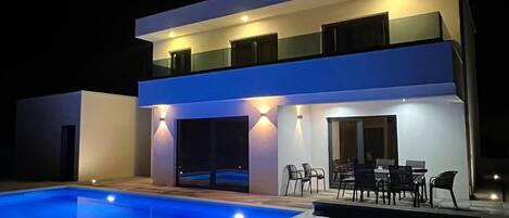 A modern two-story house with a swimming pool, outdoor seating, and a well-lit exterior.