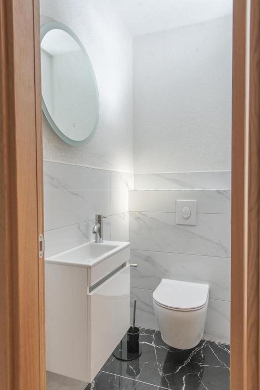 A modern bathroom with a white sink, a round mirror, a toilet, and a black marble floor.