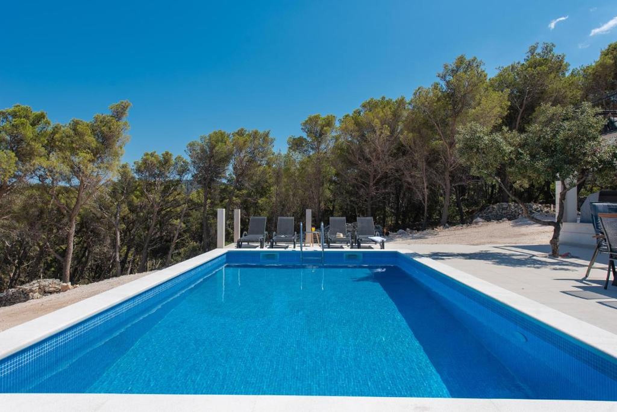A rectangular swimming pool with clear blue water, surrounded by trees and equipped with lounge chairs.