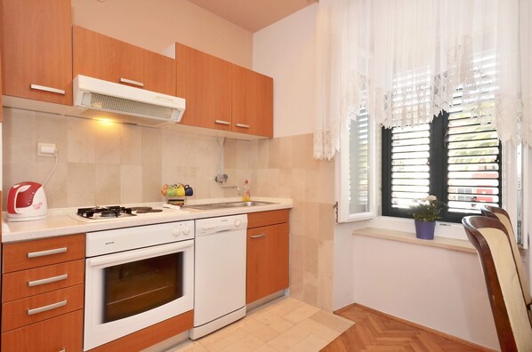 A kitchen with wooden cabinets, a gas stove, a dishwasher, and a window with blinds. - Apartment Oscar (Split)