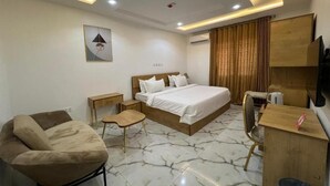 A hotel room with a bed, a desk, a chair, a small table, a wall-mounted TV, and a ceiling light fixture. - Nouveau Apartment (Abuja)