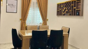 A dining area with a table, chairs, a chandelier, curtains, a wall-mounted shelf, and a framed sign. - Nouveau Apartment (Abuja)