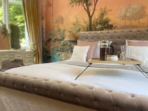 Un letto con testiera imbottita, un comodino con una lampada e un vassoio con teiera e bicchieri.