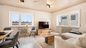 Ein modernes Wohnzimmer mit Holzboden, einem Flachbildfernseher, einem Esstisch mit Stühlen, einer Couch und einer Heizung.
