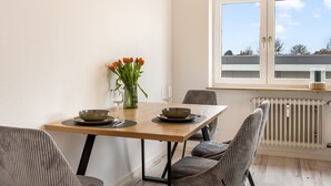 Ein Essbereich mit einem Holztisch, zwei Stühlen, einer Blumenvase und einem Fenster mit Blick auf Gebäude.