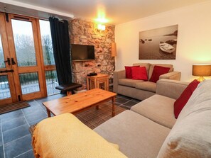 A living room with a stone wall, a flat-screen TV, a sofa, a coffee table, and a window with curtains. - Valley Cottage, Penzance (Penzance)