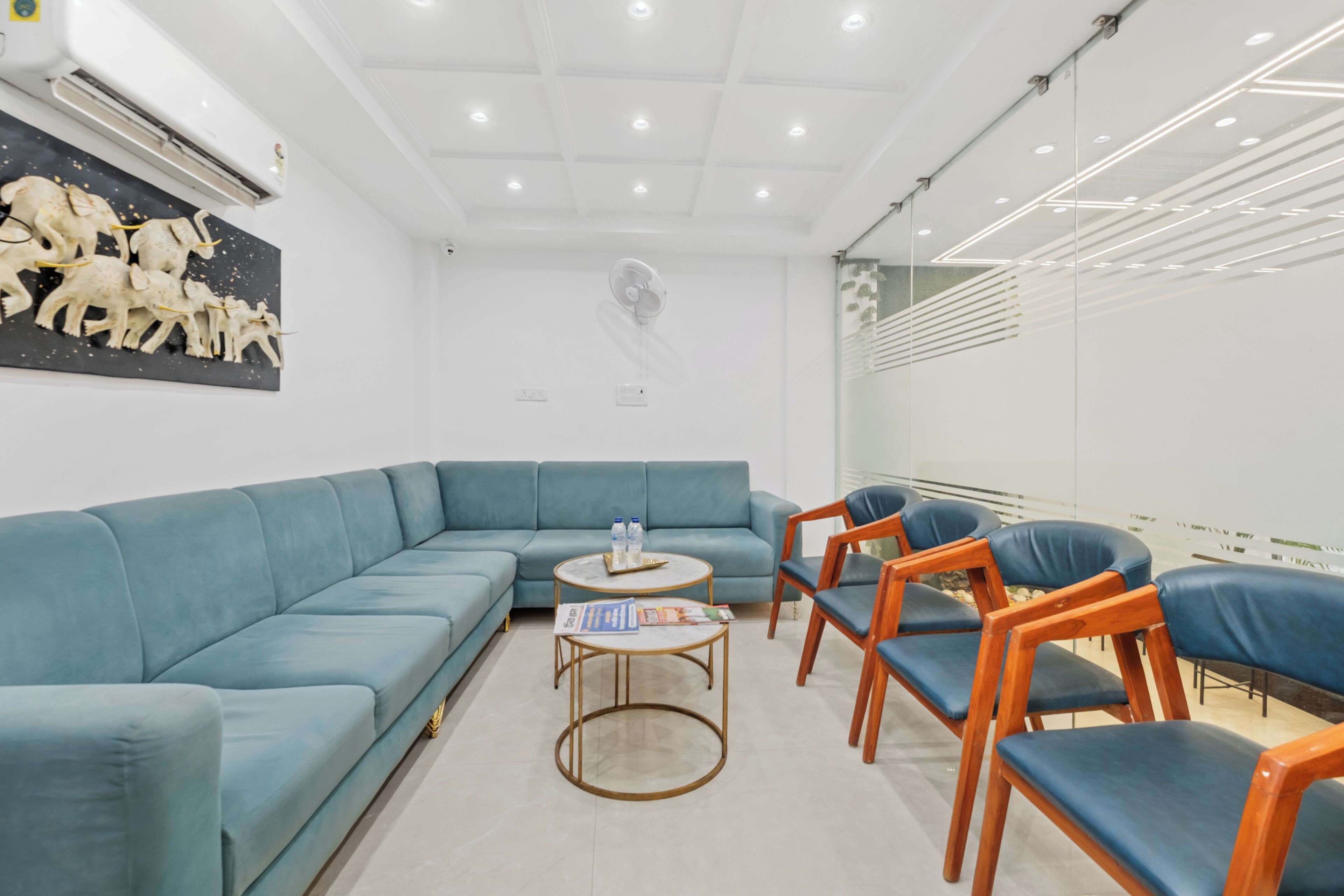 A modern room with a blue sofa, wooden chairs, a small round table with magazines, and a wall-mounted sculpture.