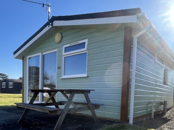 A small, single-story mobile home with a picnic table in front, a satellite dish on the roof, and a number '156' on the window.