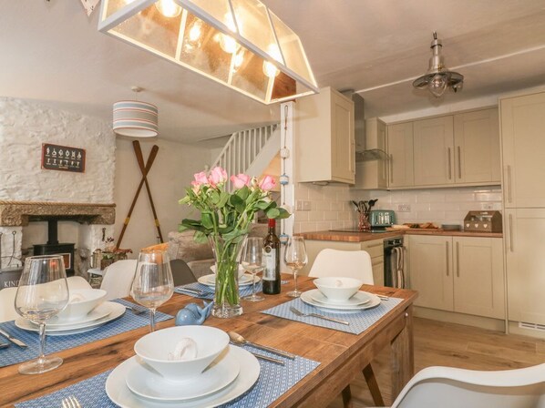 A dining area with a wooden table set for four, a vase of pink flowers, and a kitchen in the background. - 14 CLIFF STREET, open-plan,in Mevagissey (St. Austell)